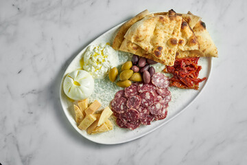 Italian antipasti snacks on white plate on marble background. Focaccia, burrata, olives, sun-dried tomatoes, salami, cheese, parmesan.