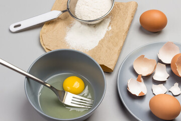 Broken egg and fork in gray bowl. Flour and sieve on paper. Chicken shells on gray plate.