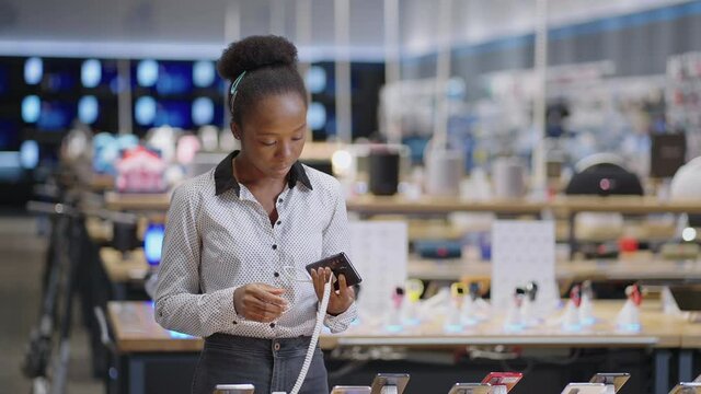 young afro-american woman is choosing cell phone, viewing exhibition samples in digital equipment store, lady is buying smartphone in retail shop