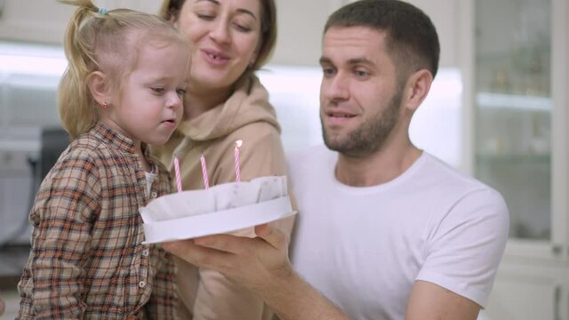 Cute Caucasian baby girl blowing candles on birthday cake with blurred parents helping at background. Cheerful daughter celebrating third birthday with father mother and sister at home. Joy childhood