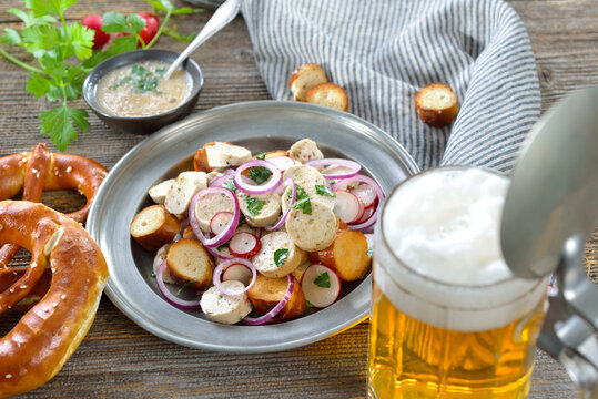 Bayerischer Wei&szlig;wurstsalat mit ger&ouml;steten Brezenscheiben auf Zinnteller dazu ein helles Bier &ndash; Delicious Bavarian salad with cut veal sausages and roasted pretzel slices with a mug of lager beer 