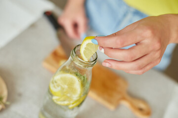 Female hand put lemon in glass bottle, making detox healthy water with lemon and rosemary