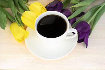 On a wooden table among two bouquets of tulips of different colors there is a white cup of coffee.