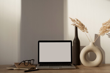 Aesthetic home office desk workspace with sunlight shadows on the wall. Blank screen laptop computer with copy space. Glasses, pampas grass in stylish vase on wooden table. Influencer lifestyle blog