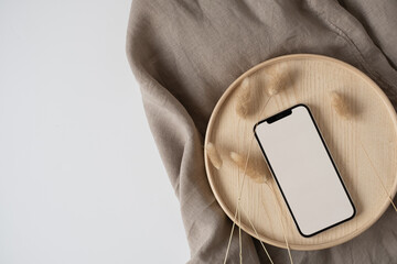 Blank screen mobile phone, rabbit tail grass on wooden tray with crumpled linen cloth blanket. Flat...