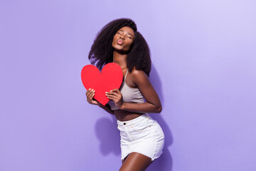 Portrait of attractive dreamy amorous girl holding in hands heart card posing sending air kiss isolated over violet purple color background