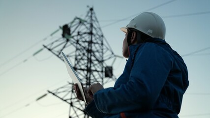 Naklejka na ściany i meble Woman power engineer in white helmet checks power line using computer on tablet, remote control of power system. High voltage electrical lines at sunset. Distribution and supply of electricity. Energy