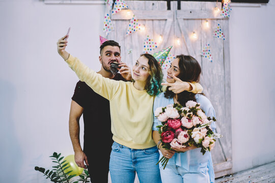 Group of young best friends posing for selfie on smartphone camera having fun together on anniversary celebration, birthday girl with flowers taking picture via telephone with guests of party