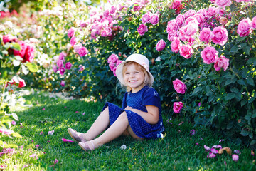 Fototapeta premium Portrait of little toddler girl in blossoming rose garden. Cute beautiful lovely child having fun with roses and flowers in a park on summer sunny day. Happy smiling baby.