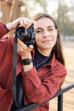 Portrait of a young photographer woman looking in the camera and having a cute smile with a vintage mirorless camera in her hands outside in the park