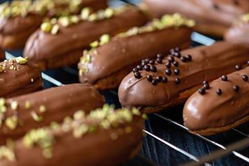 French dessert eclairs or profiteroles with milk chocolate glaze, drops and pistachios on a baking rack. Pastry cakes with cream and topping. Soft selective focus.
