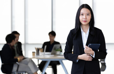 Young Asian businesswoman in black suit standing and holding tablet  computer posing to camera in office with her team talking in blur background