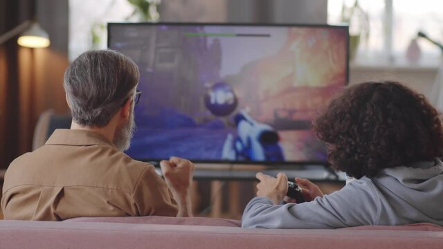 Back view of grey haired father and young teen son sitting on sofa at home and playing shooter video game on TV