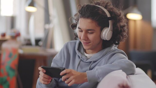 Cheerful teenage boy in wireless headphones sitting on sofa at home, smiling and playing game on smartphone