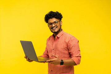 Indian male manager, company worker holding laptop in hand and smiling looking at camera isolated on yellow background with place for text