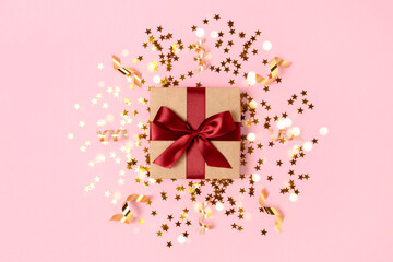 Gift box tied with red ribbon on a pink background with gold stars confetti. Festive composition.