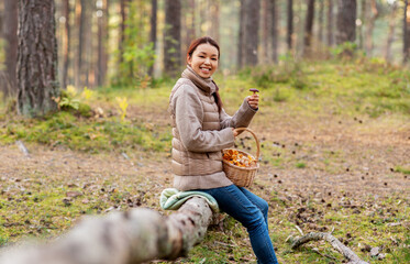 Naklejka na ściany i meble picking season, leisure and people concept - young asian woman with mushrooms in basket in autumn forest