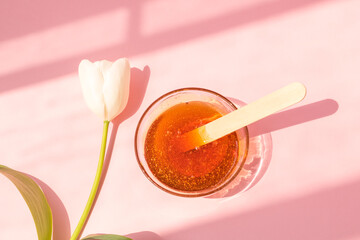 Shugaring paste in a glass bowl on a pink background. Hair removal, epilation, wooden spatula for sugaring paste, tulip.