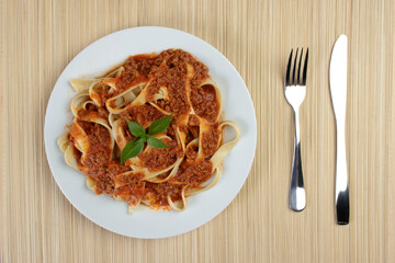 Pasta with bolognese sauce and basil leaves served on a plate on a bamboo mat. Fork and knife for the meal. Top view photo.