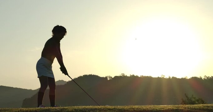 Slow Motion Golf Swing Drive Shot. Silhouette Asian female golfer enjoying vacation luxury resort using driver to tee off golf course fairway over sunset background.