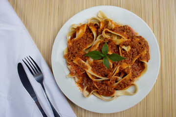 Pasta with bolognese tomato sauce and basil leaves served on a white plate in the background, bamboo mat and napkin with fork and knife beside it. top view photo.