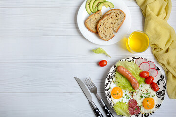Plate with scrambled eggs, sausages, fresh tomatoes and salad, avocado on a light wooden background, top view. Layout, copy space. Delicious and nutritious breakfast
