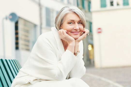 Outdoor portrait of beautiful and elegant middle age 55 - 60 year old woman, wearing white jacket, sitting on bench outside