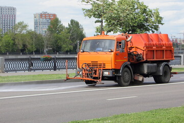 New orange watering machine street cleaner truck spraying dry asphalt road, street washing in Moscow, city improvement by municipal services on a sunny summer day