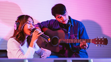 Obraz premium Pretty Asian female singer recording songs by using a studio microphone with male playing guitar in blue and red light. Performance and show in the music business. Duet session.