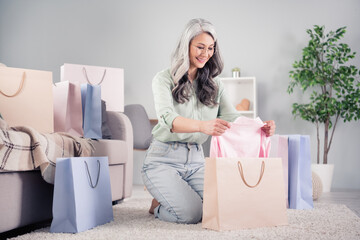 Full body portrait of cheerful aged person it on knees hands taking clothing from bag toothy smile indoors