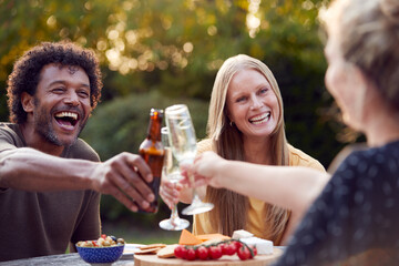 Group Of Friends Celebrating With Beer And Champagne As They Sit At Table In Garden With Snacks