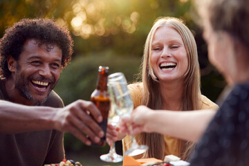 Group Of Friends Celebrating With Beer And Champagne As They Sit At Table In Garden With Snacks