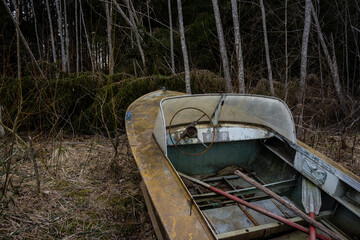 An old rusty metal boat stands on the shore between trees with a rudder and paddles criss-crossed into the boat