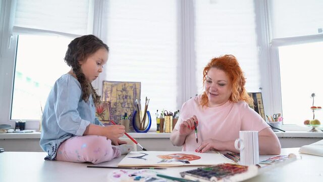 Happy family art leisure together concept. Mother and daughter painting together at home with paintbrushes and watercolors. Young woman sitting and helping or teaching daughter to draw. 4k footage