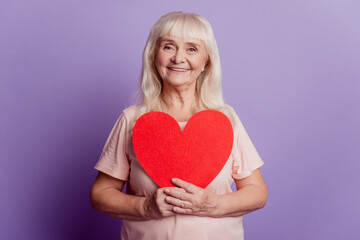 Positive elderly woman hold big valentine heart card isolated on violet background