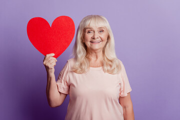 Photo of smiling old pretty woman hold big red card heart isolated on violet background - Powered by Adobe