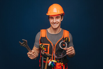 Fototapeta premium Smiling builder in a helmet is holding glasses and wrench, isolated over blue background