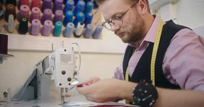 Tailor seated at his workbench sewing a bespoke garment on a sewing machine with sample colour yarns in the background mounted on the wall