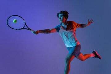 Young caucasian man playing tennis isolated on purple-blue studio background in neon, action and...