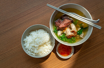 Top view Asian meal, a bowl of pig's blood soup and pork with steam white rice and spicy-sour sauce, natural light on table. Pig's blood soup and pork is called Tom Leard Moo in the Thailand language.