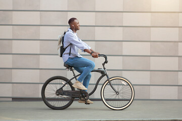 Side view of cool black guy with earphones racing on his bicycle near brick wall downtown