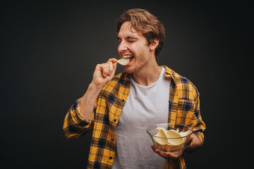 Young blond man in yellow shirt is eating chips and holding plate with it, isolated over black background