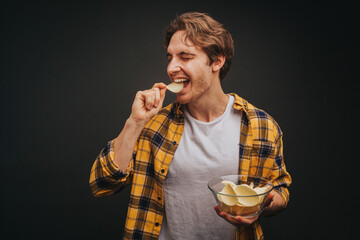 Young blond man in yellow shirt is eating chips and holding plate with it, isolated over black...