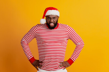 Fototapeta na wymiar Portrait of angry guy in santa hat hands hips isolated on yellow background