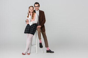 man in eyeglasses hugging retro style woman while sitting on high chair on grey background