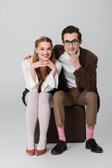 joyful couple in retro outfit smiling at camera while sitting on suitcases on grey background