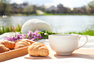 Summer picnic near the river with cup of coffee and croissants. Selective focus. Outdoor lunch or breakfast. Summer mood
