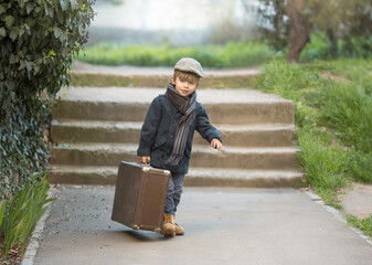 a little boy is carrying a big suitcase on the street, he is going on a trip. He's wearing trendy vintage clothes and rough boots