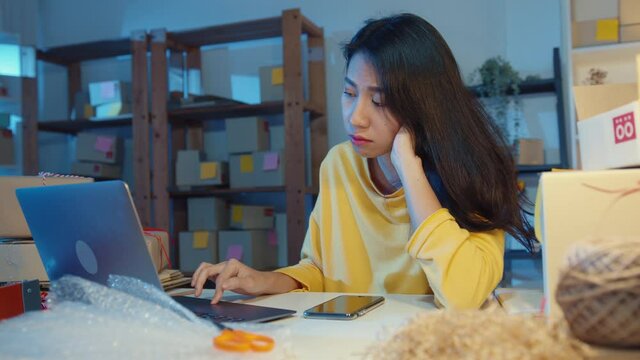 Young Asia Businesswoman Look Around Room Full Of Product Stuff And Parcel Box Feel Stress And Upset With Bad Sell In Home Office At Night. Small Business Owner, Online Market Delivery Concept.