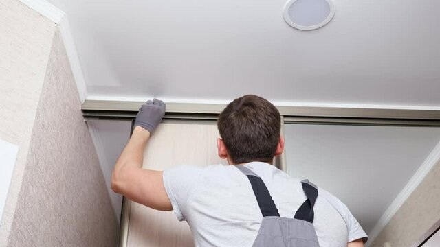 Employee furniture assembler in white t-shirt and jumpsuit checks wooden sliding door of modern wardrobe with mirror close back low angle shot